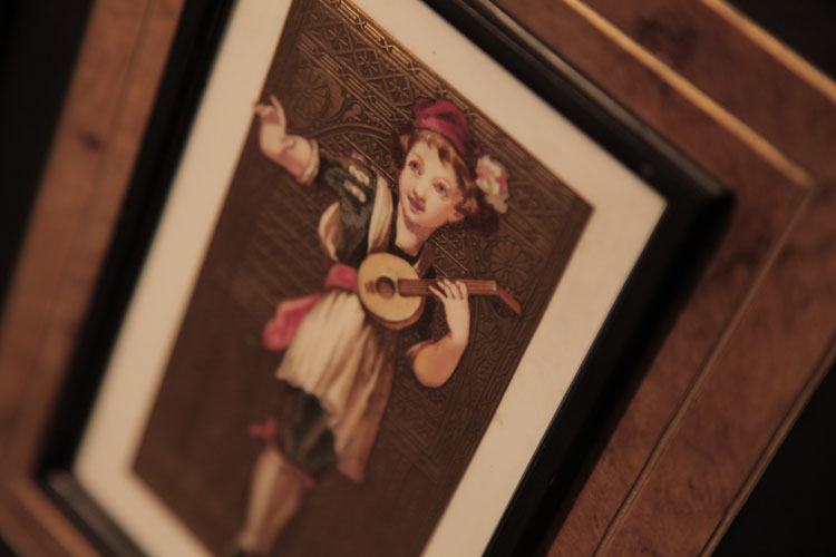 Detail of the hand-painted ceramic tile mounted on piano cabinet edged with a black border and gold embossed, patterned background. Tile depicts a young boy playing a lute  