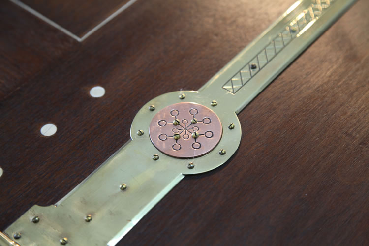 Arts and Crafts objects were simple in form, without superfluous or excessive decoration, and how they were constructed was often still visible. Here the hinges, often hidden in modern furniture, are a strong element of the piano's design. The hinges are large and stamped with a circular design. Returning to Norse mythology, this symbol appears to depict the nine worlds, a symbol of Norse cosmology. 